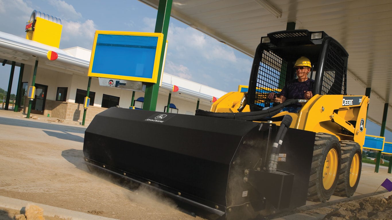 Chargeuse à direction différentielle John Deere équipée de l’accessoire balai ramasseur qui balaie